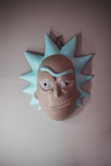A stylized mask resembling a cartoon character with spiky blue hair and exaggerated facial features, including large eyes and a prominent brow. The mask is mounted on a plain wall and conveys an abstract and humorous aesthetic.