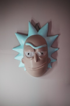 A stylized mask resembling a cartoon character with spiky blue hair and exaggerated facial features, including large eyes and a prominent brow. The mask is mounted on a plain wall and conveys an abstract and humorous aesthetic.