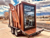 A small modern, rectangular structure is built on a trailer, with large glass windows and a wooden exterior. A tall, orange flag with the word 'Milieu' waves beside it. The background features a rural setting with railway tracks, dry vegetation, and mountains under a partly cloudy sky.