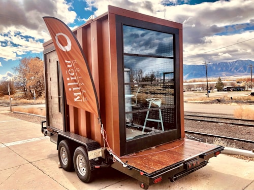A small modern, rectangular structure is built on a trailer, with large glass windows and a wooden exterior. A tall, orange flag with the word 'Milieu' waves beside it. The background features a rural setting with railway tracks, dry vegetation, and mountains under a partly cloudy sky.