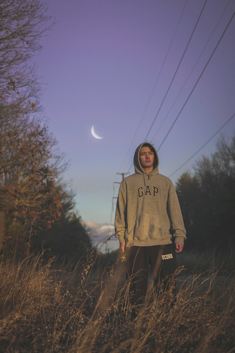 man wearing gray GAP hooded jacket standing near road surrounded with green trees showing crescent moon