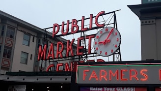 A large neon sign displaying the words 'PUBLIC MARKET CENTER' with a prominent round analog clock positioned above it. The clock has red numbers and hands. Below the main sign, there is another sign that reads 'FARMERS' in red neon. The background includes parts of adjacent buildings with windows and architectural details.