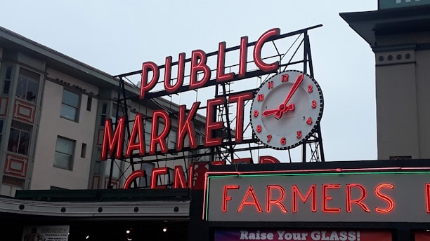 A large neon sign displaying the words 'PUBLIC MARKET CENTER' with a prominent round analog clock positioned above it. The clock has red numbers and hands. Below the main sign, there is another sign that reads 'FARMERS' in red neon. The background includes parts of adjacent buildings with windows and architectural details.