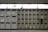 A wall of lockers, some with visible wear and tear, arranged in a grid pattern. Each locker has a numbered label and a small display panel. Above the lockers, there's an illuminated emergency exit sign with a running figure and an arrow. The lighting is bright, and the area appears clean and organized.
