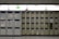 A wall of lockers, some with visible wear and tear, arranged in a grid pattern. Each locker has a numbered label and a small display panel. Above the lockers, there's an illuminated emergency exit sign with a running figure and an arrow. The lighting is bright, and the area appears clean and organized.