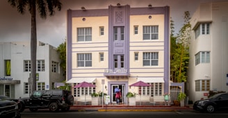 A three-story building with a pastel facade featuring vertical lines and decorative details. The sign reads 'Hotel Shelley' above the entrance. Two purple umbrellas are set up on a patio with plants and a white railing in front. A person in a red top and another in blue are visible near the entrance. Palm trees and parked vehicles line the street.