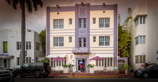 A three-story building with a pastel facade featuring vertical lines and decorative details. The sign reads 'Hotel Shelley' above the entrance. Two purple umbrellas are set up on a patio with plants and a white railing in front. A person in a red top and another in blue are visible near the entrance. Palm trees and parked vehicles line the street.