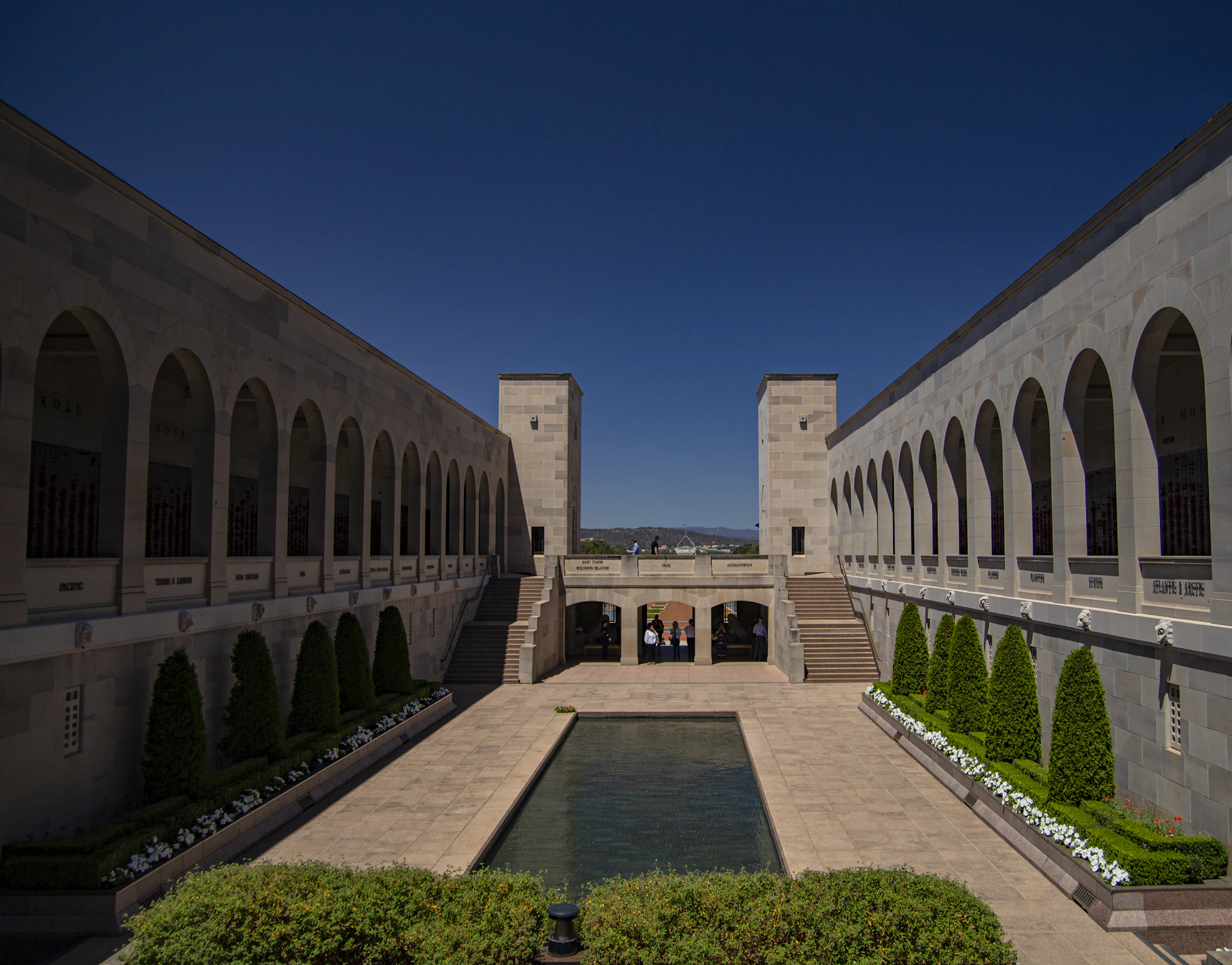A symmetrical courtyard surrounded by stone walls with archways on each side. In the center is a reflective pool bordered by neatly trimmed bushes and small flowering plants. Stairs on either side lead up to a balcony, and a deep blue sky provides a contrasting backdrop.
