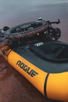 A yellow inflatable raft with the brand name 'Rogue' is positioned on a wet, muddy surface. A bicycle appears to be mounted or attached on top of the raft, with visible knobby tires suggesting it might be used for rough terrain.