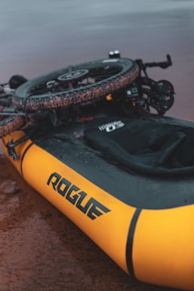 A yellow inflatable raft with the brand name 'Rogue' is positioned on a wet, muddy surface. A bicycle appears to be mounted or attached on top of the raft, with visible knobby tires suggesting it might be used for rough terrain.