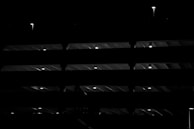 A multi-level structure illuminated by small lights at regular intervals, creating a pattern of light and shadow. The beams and floors of the structure are silhouetted against the dark background, giving a sense of depth and architecture.