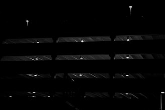 A multi-level structure illuminated by small lights at regular intervals, creating a pattern of light and shadow. The beams and floors of the structure are silhouetted against the dark background, giving a sense of depth and architecture.
