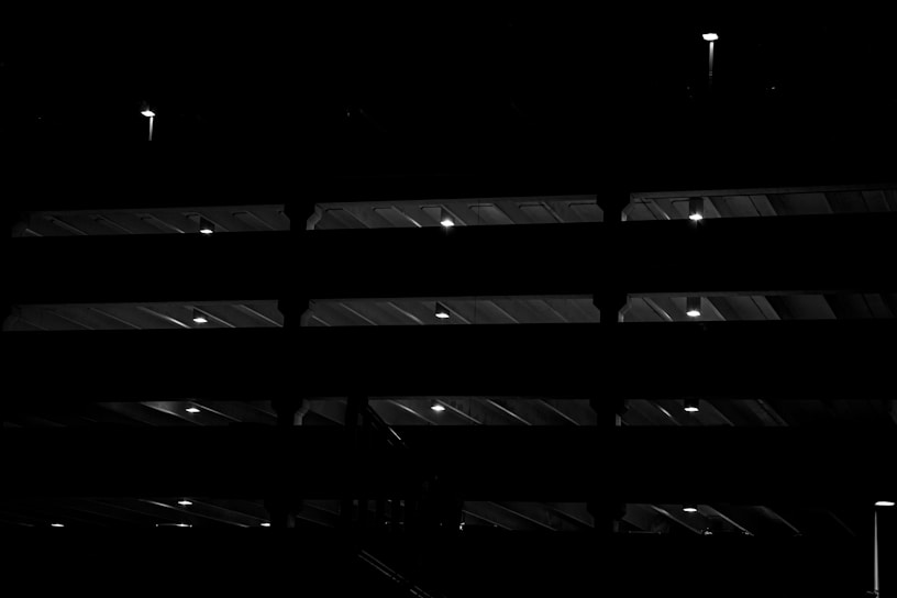 A multi-level structure illuminated by small lights at regular intervals, creating a pattern of light and shadow. The beams and floors of the structure are silhouetted against the dark background, giving a sense of depth and architecture.