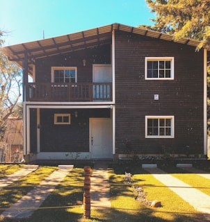 A two-story wooden house with a sloped roof and a small balcony on the upper floor. The facade features wooden paneling with white trim around the windows and doors. It is surrounded by well-maintained green lawns, paved paths, and some trees, under a clear blue sky.