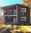A two-story wooden house with a sloped roof and a small balcony on the upper floor. The facade features wooden paneling with white trim around the windows and doors. It is surrounded by well-maintained green lawns, paved paths, and some trees, under a clear blue sky.