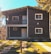 A two-story wooden house with a sloped roof and a small balcony on the upper floor. The facade features wooden paneling with white trim around the windows and doors. It is surrounded by well-maintained green lawns, paved paths, and some trees, under a clear blue sky.