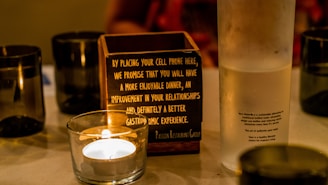 A table displays a lit candle in a glass holder, a wooden box with writing about the benefits of placing a cell phone inside, and a frosted water bottle with printed text describing the sustainable qualities of the water inside. Black cups are also arranged around the table.