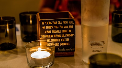 A table displays a lit candle in a glass holder, a wooden box with writing about the benefits of placing a cell phone inside, and a frosted water bottle with printed text describing the sustainable qualities of the water inside. Black cups are also arranged around the table.