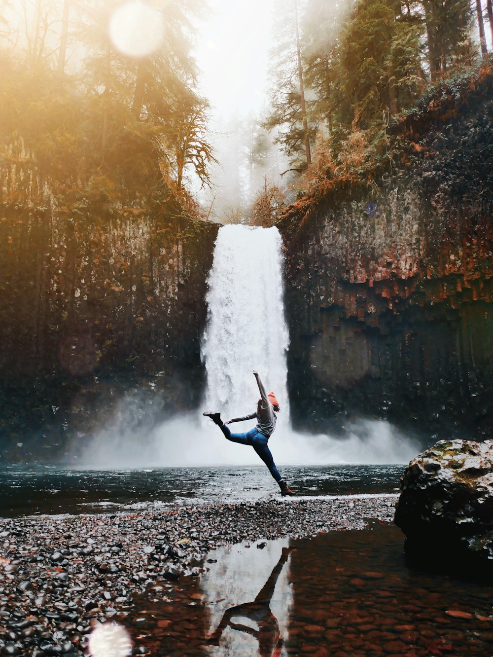 woman jumping in front of waterfalls during daytime