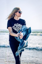 A young woman with long hair and sunglasses holds a denim jacket while standing at the beach. The waves gently crash in the background and she wears a black shirt with the phrase 'Born to Worship'. The atmosphere is sunny and breezy.