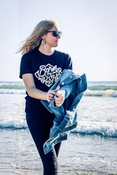 A young woman with long hair and sunglasses holds a denim jacket while standing at the beach. The waves gently crash in the background and she wears a black shirt with the phrase 'Born to Worship'. The atmosphere is sunny and breezy.