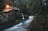 A rustic wooden cabin is situated in a dense forest, illuminated by warm light emanating from its windows. A small waterfall flows beside the cabin, with a serene stream winding through the lush greenery. Moss-covered tree branches frame the scene, adding to the natural ambiance.