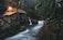 A rustic wooden cabin is situated in a dense forest, illuminated by warm light emanating from its windows. A small waterfall flows beside the cabin, with a serene stream winding through the lush greenery. Moss-covered tree branches frame the scene, adding to the natural ambiance.