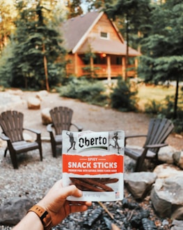 A person holds a packet of Oberto spicy snack sticks in the foreground. In the background, there's a cozy cabin surrounded by trees, with several Adirondack chairs arranged around a fire pit area. The setting appears to be a wooded area, providing a rustic and serene atmosphere.