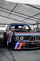 A classic race car with a sleek black body adorned with vibrant blue, red, and white racing stripes. The setting appears to be a covered area, possibly a garage or pit stop, with the vehicle prominently positioned. The front of the car displays four large headlights and the brand logo. The words 'RECARO' and 'TOW' are visible, indicating racing features.