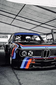 A classic race car with a sleek black body adorned with vibrant blue, red, and white racing stripes. The setting appears to be a covered area, possibly a garage or pit stop, with the vehicle prominently positioned. The front of the car displays four large headlights and the brand logo. The words 'RECARO' and 'TOW' are visible, indicating racing features.