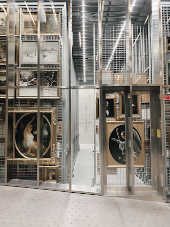 A storage facility with metal racks containing numerous framed paintings, some of which depict scenes and figures. The environment is well-lit with rows of fluorescent lights and the floor has a metallic, industrial appearance.