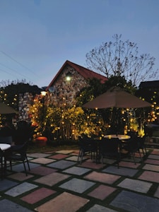 A cozy outdoor seating area is adorned with string lights, surrounding stone pavement, and vibrant greenery under an early evening sky. A small stone house with a triangular roof serves as a backdrop, with chairs and tables scattered around the patio.