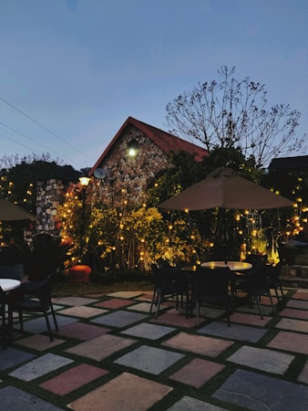 A cozy outdoor seating area is adorned with string lights, surrounding stone pavement, and vibrant greenery under an early evening sky. A small stone house with a triangular roof serves as a backdrop, with chairs and tables scattered around the patio.