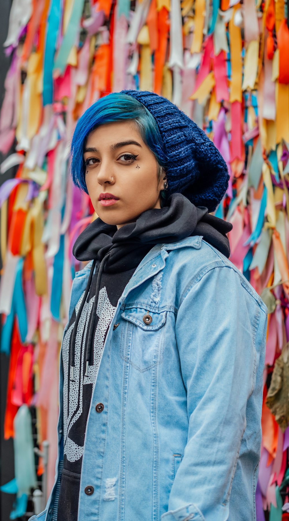 woman with blue hair wearing blue denim jacket standing beside wall with bows