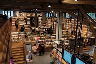 An expansive bookstore with multiple levels and wooden bookshelves filled with a variety of books. People are browsing and walking around the store. The space is well-lit with overhead lights, and there are some plants placed strategically to enhance the atmosphere.