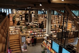 An expansive bookstore with multiple levels and wooden bookshelves filled with a variety of books. People are browsing and walking around the store. The space is well-lit with overhead lights, and there are some plants placed strategically to enhance the atmosphere.