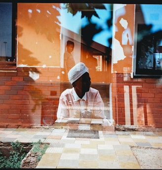 A double-exposure photograph features a person wearing a white turban and a light-colored shirt, overlaid with an urban scene that includes an orange and red brick wall. The same image reveals another figure, a child, partially visible in the background, with elements such as a window and patterns on the ground being discernible.