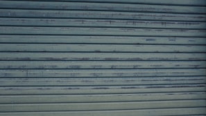 A close-up view of a metallic, corrugated garage door with a weathered appearance and horizontal lines. It has a grayish-blue color and shows signs of rust and wear.