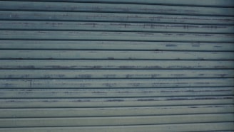 A close-up view of a metallic, corrugated garage door with a weathered appearance and horizontal lines. It has a grayish-blue color and shows signs of rust and wear.