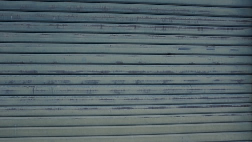A close-up view of a metallic, corrugated garage door with a weathered appearance and horizontal lines. It has a grayish-blue color and shows signs of rust and wear.