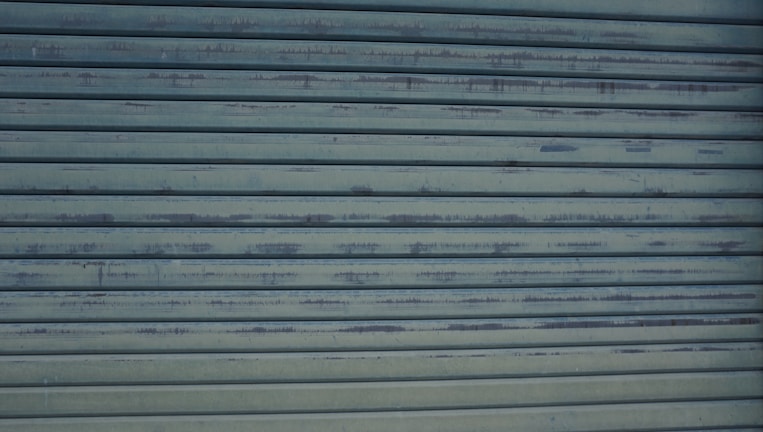 A close-up view of a metallic, corrugated garage door with a weathered appearance and horizontal lines. It has a grayish-blue color and shows signs of rust and wear.