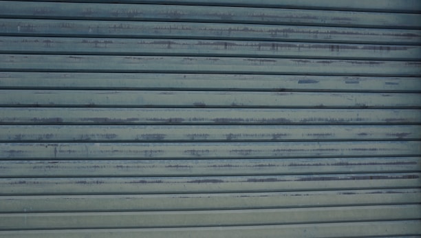 A close-up view of a metallic, corrugated garage door with a weathered appearance and horizontal lines. It has a grayish-blue color and shows signs of rust and wear.