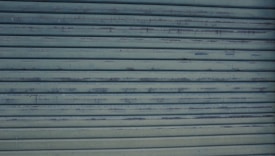 A close-up view of a metallic, corrugated garage door with a weathered appearance and horizontal lines. It has a grayish-blue color and shows signs of rust and wear.
