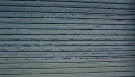 A close-up view of a metallic, corrugated garage door with a weathered appearance and horizontal lines. It has a grayish-blue color and shows signs of rust and wear.