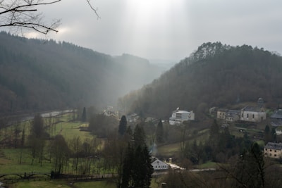 A rural village nestled in a valley surrounded by densely forested hills. Soft light filters through the clouds, casting a serene glow over the landscape. The village features a cluster of homes, a church, and open grassy fields. A small river winds through the valley, bordered by bare trees.