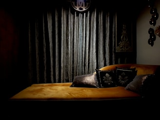A dimly lit room features a luxurious, mustard-colored velvet chaise lounge adorned with decorative dark pillows. Behind the lounge is a backdrop of heavy, dark curtains, complemented by ornate wall decorations and a round, abstract symbol hanging above. The overall ambiance suggests a sense of vintage elegance.