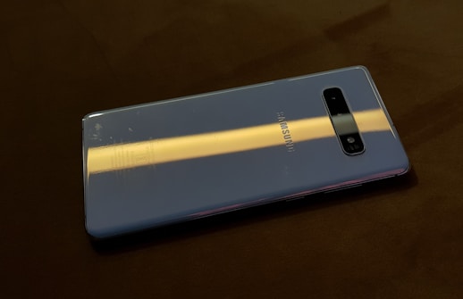 A smartphone placed on a dark surface, displaying a reflective back cover with a slight glare from lighting. The brand and camera lenses are visible on the back of the phone.