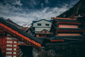 Traditional buildings with ornate architectural details are set against a backdrop of snow-capped mountains and a partly cloudy sky. The structures feature dark and burnt orange colors, with intricate woodwork and roof designs. Tall, dense trees line the mountainside, contributing to a serene, natural setting.