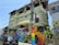 A residential building with a green facade is decorated with a colorful mural on a wall and a rainbow flag hanging on the balcony. The street in front features a black car and numerous power lines crisscrossing above. The mural includes intricate patterns in vibrant colors, and a utility pole is painted with rainbow stripes.