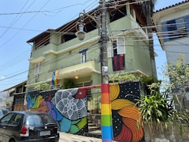 A residential building with a green facade is decorated with a colorful mural on a wall and a rainbow flag hanging on the balcony. The street in front features a black car and numerous power lines crisscrossing above. The mural includes intricate patterns in vibrant colors, and a utility pole is painted with rainbow stripes.
